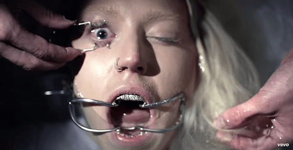Her mouth and one eye forced open using sadistic surgical methods. We later see her forced to ingest a liquid and choking on it. These images are flashed extremely quickly.